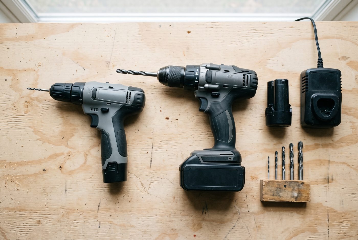 A compact cordless drill and a full-size cordless drill side by side on plywood, with a spare battery, a charger and a few drill bits.
