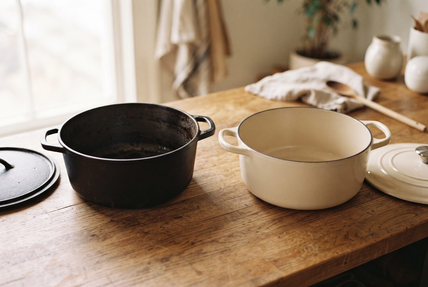 A bare black cast iron Dutch oven next to a cream enameled cast iron Dutch oven on a wooden counter, lids off.