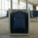 A compact navy softside carry-on spinner suitcase standing inside a metal airline bag sizer at an airport gate, wheels included inside the frame.