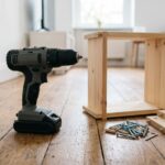 A gray and black cordless drill with its battery attached, standing on a wooden floor beside a half-assembled flat-pack bookshelf and a pile of screws.