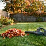 A compact cordless leaf blower with its battery clipped in, resting on a small backyard lawn beside a neat pile of red and orange maple leaves, with a wooden fence and flower beds behind.