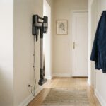 A cordless stick vacuum hanging on a wall-mounted charging dock in a small apartment hallway, its cable running down to an outlet.