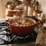 A red enameled cast iron Dutch oven simmering beef and vegetable stew on a gas stovetop, lid tilted to one side, wooden spoon on a linen towel.