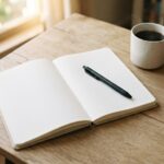 An open planner with blank ruled pages and a black pen on a wooden desk, beside a cup of coffee and a small succulent in morning light.