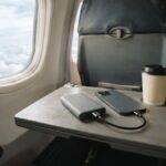 A compact gray power bank charging a smartphone by a short cable on an airplane tray table, next to a paper coffee cup and the window.