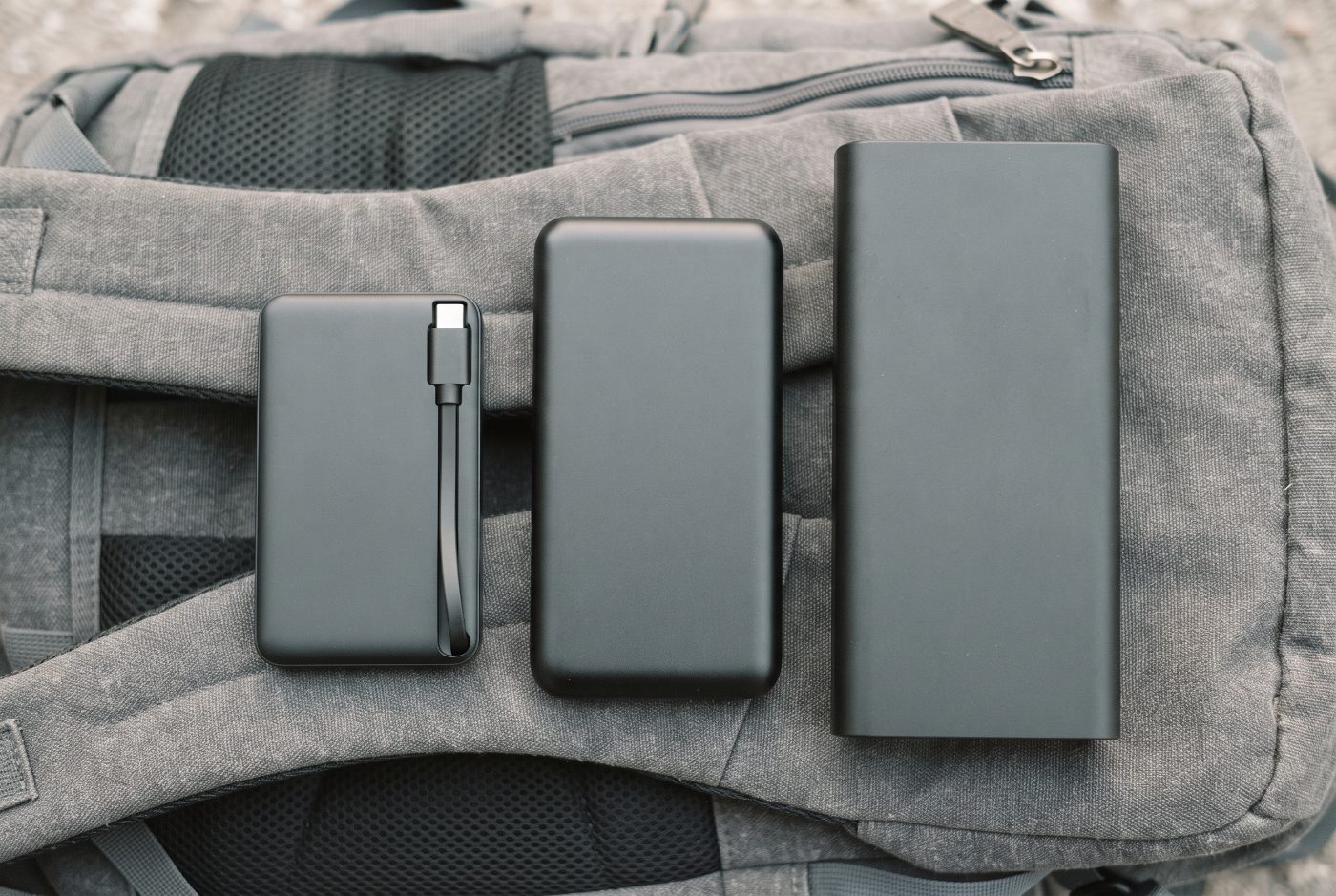 Three gray power banks side by side on a travel backpack, from a slim one with a built-in cable to a large brick.